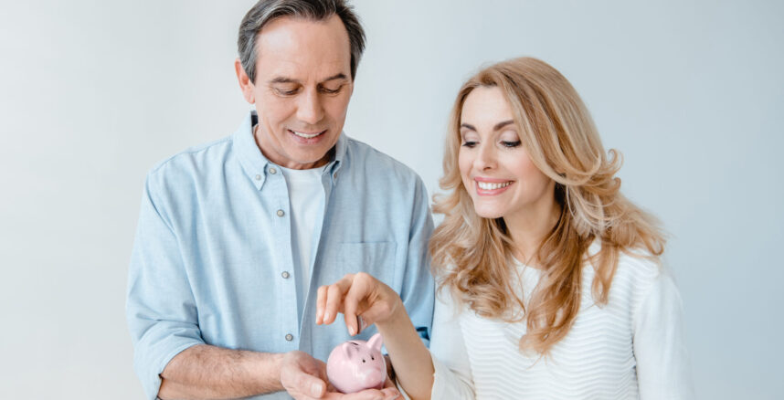 Smiling middle aged couple putting coin into small piggy bank isolated on grey pedro braggio financas a dois scaled